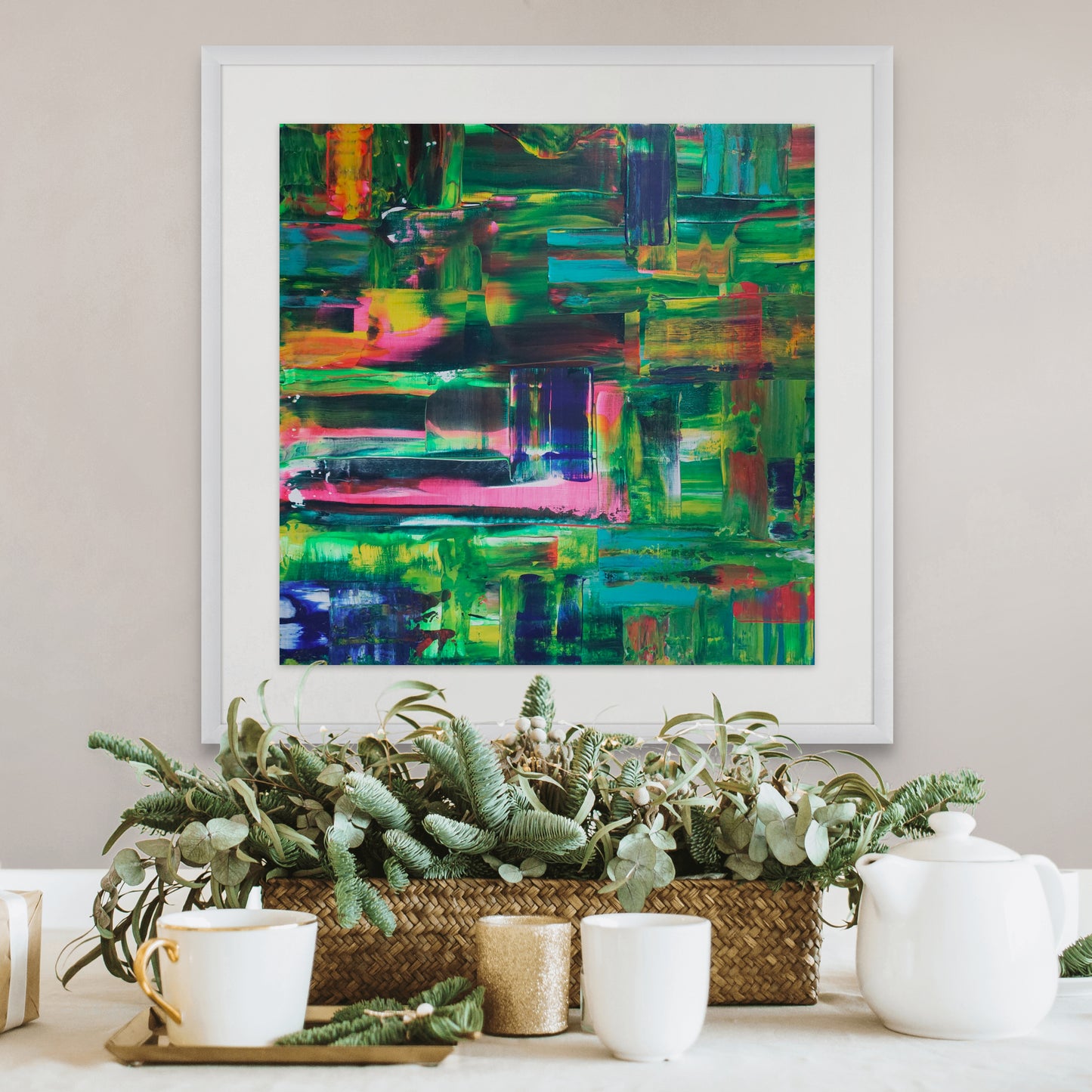 Colorful abstract painting in a white frame above a decorative table setting with plants and teacups.