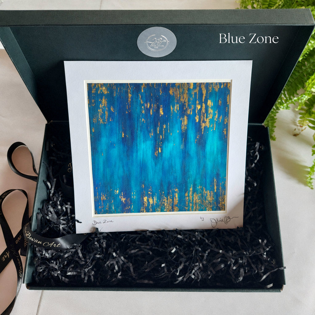 An abstract art print featuring textured blue and teal paint with gold leaf-like accents, presented in a white mat within an open black gift box with black paper shred.
