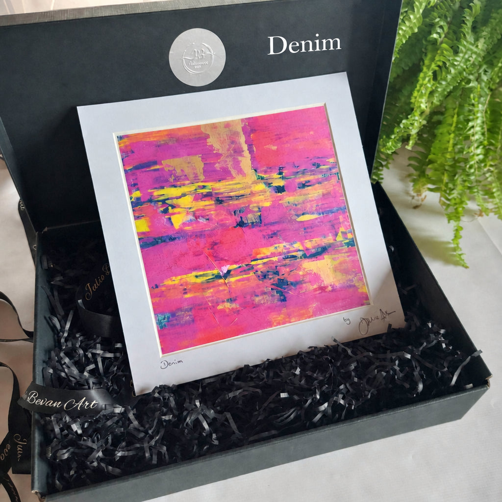 A framed abstract art print with vibrant pink, purple, and yellow brushstrokes, presented inside a dark gray gift box with black shredded paper filler. The print has a wide white border and is signed by the artist, with a green fern visible in the background.