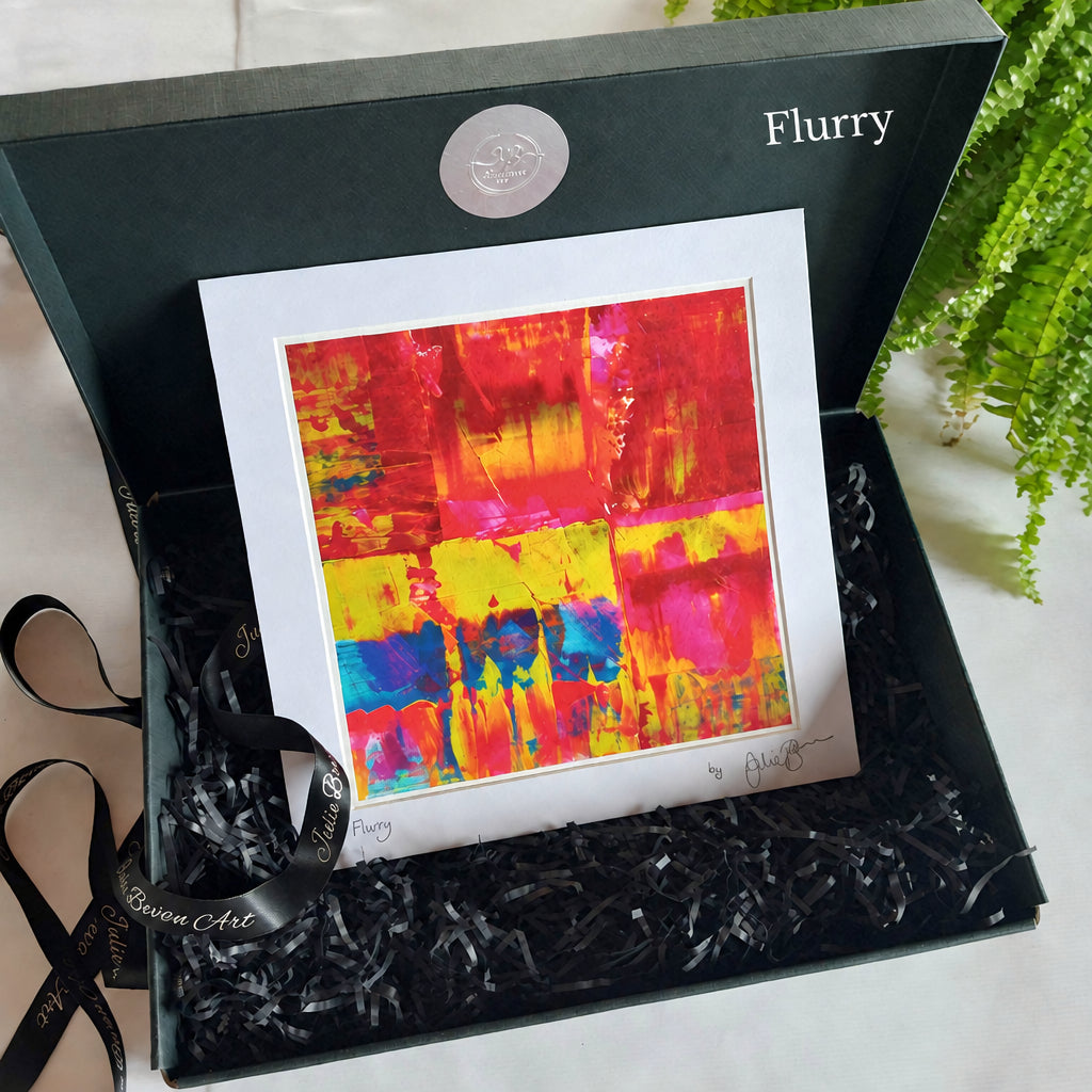 An abstract colorful art print with red, yellow, pink, and blue brushstrokes, matted in white and presented inside a dark green gift box with black paper shred and a black ribbon.