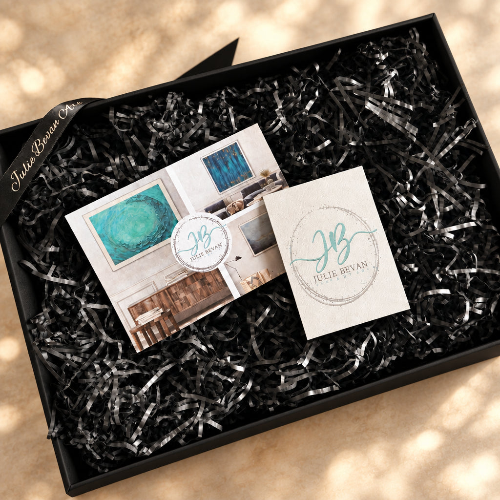 A black gift box filled with black shredded paper packaging, containing two cards: one printed with interior design photos and a round monogram seal, and one white card with a teal circular 