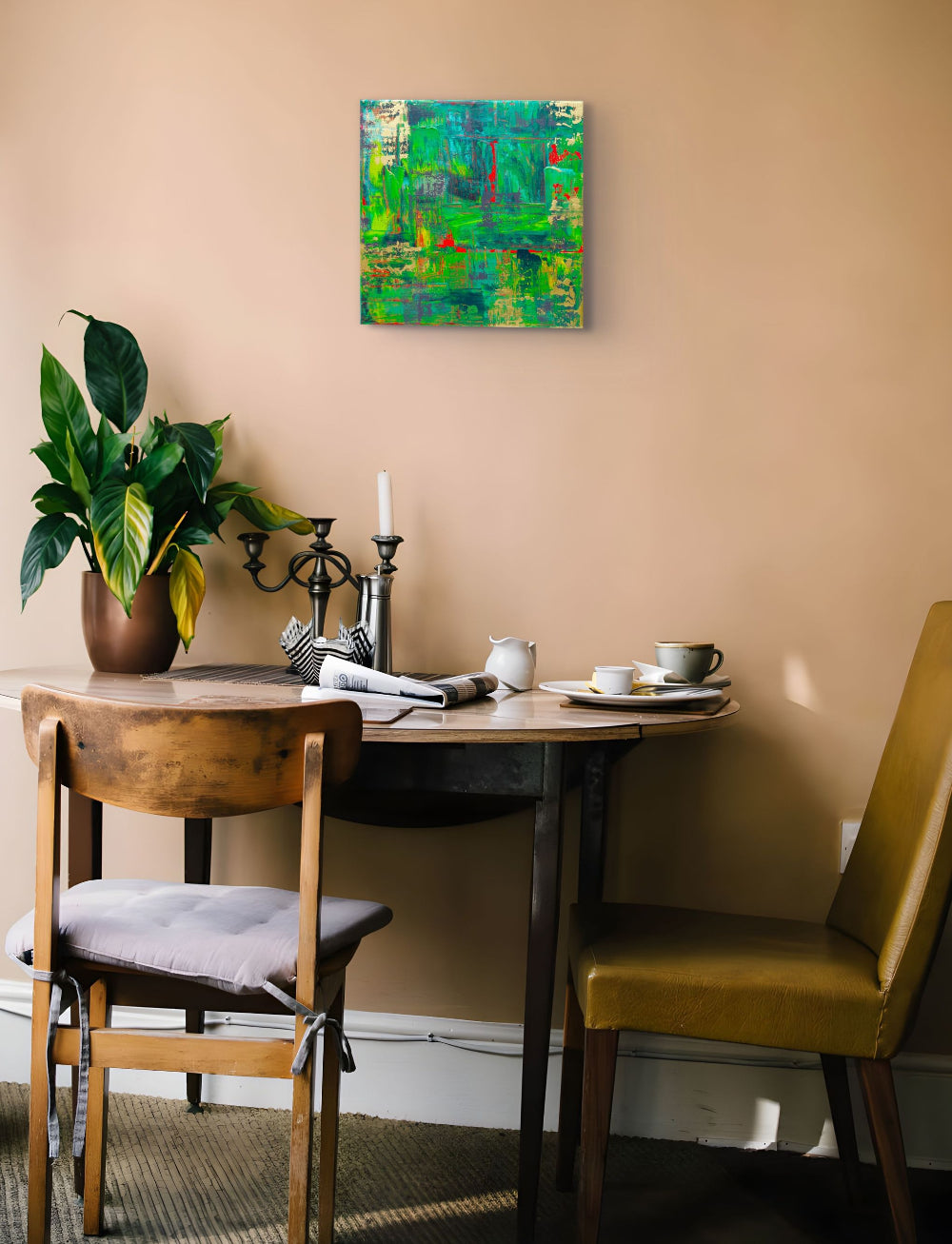 A square abstract painting with vibrant green, blue, and red brushstrokes hangs on a warm beige wall above a wooden dining table with chairs, a potted plant, and tableware. Samhain no. 3 limited edition print by Julie Bevan