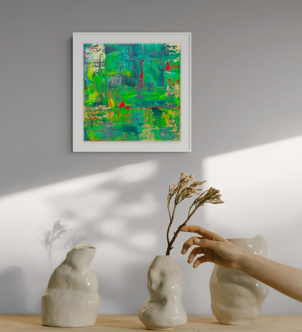 A framed abstract painting with vibrant green, teal, and red brushstrokes hangs on a light gray wall. Below, a wooden surface holds three sculptural off-white ceramic vases, with a hand placing a dried brown twig into the center vase. Samhain no. 3 limited edition print by Julie Bevan