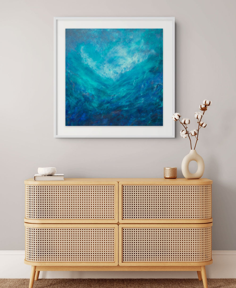A framed abstract painting with swirling blue and teal waves hangs on a light gray wall above a light wood cane-topped sideboard with decorative objects.