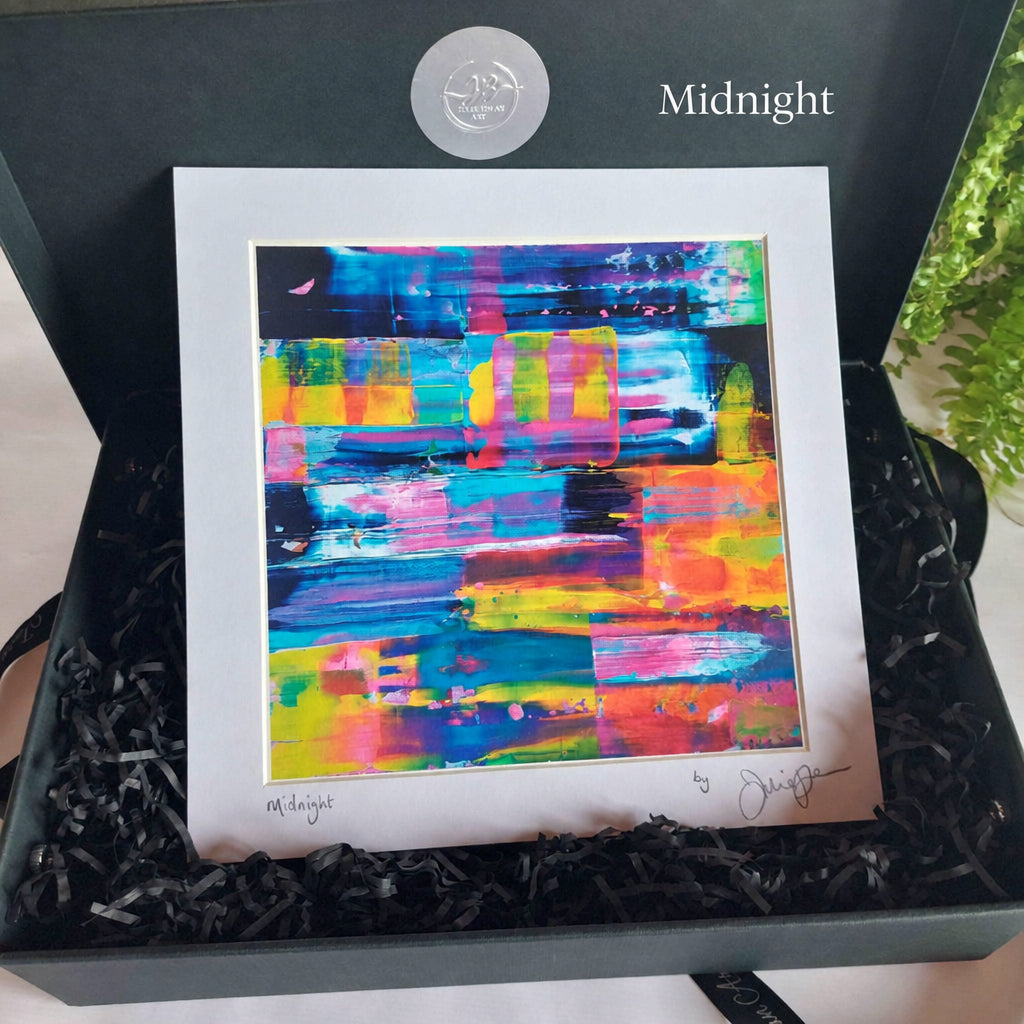 A colorful abstract art print with a wide white mat board, presented inside a dark gray gift box filled with black shredded paper. The artwork features a vibrant mix of blue, pink, yellow, green, and orange brushstrokes, with a silver circular sticker on the inside lid and an artist signature at the bottom right.