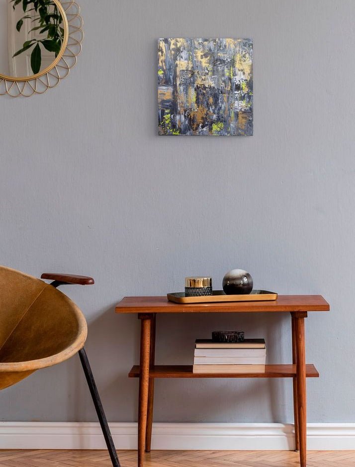 A square abstract canvas print with blue, gray, gold, and white brushstrokes, displayed on a light gray wall above a wooden side table in a modern room setting. Samhain no. 4 original abstract canvas painting by Julie Bevan