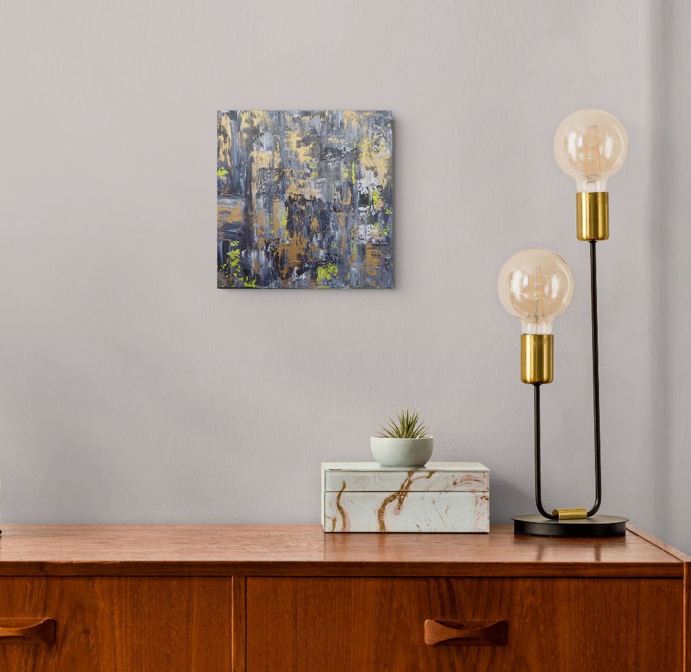 A square abstract canvas painting with blue, gray, gold, and white brushstrokes hangs on a light gray wall above a wooden cabinet. A small potted succulent sits on a white marble box on the cabinet, and a black two-light table lamp with gold accents stands to the right. Samhain no. 4 original abstract canvas painting by Julie Bevan