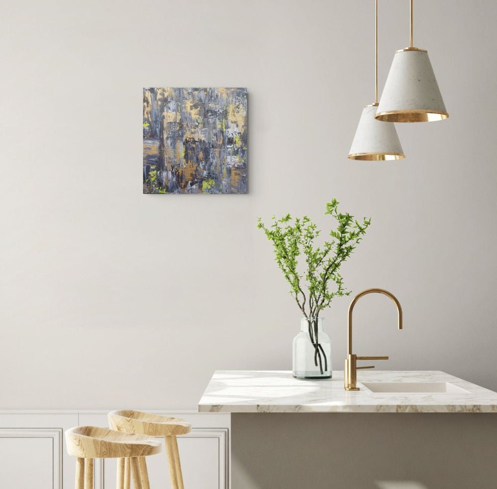 An abstract canvas painting with blue, gray, and gold tones hangs on a light gray wall above a kitchen island with a white marble countertop, gold faucet, and a vase of green branches. Samhain no. 4 original abstract canvas painting by Julie Bevan