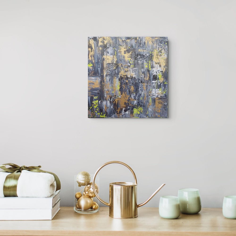 An abstract canvas painting with textured brushstrokes in gray, gold, and blue hangs on a light gray wall above a wooden shelf styled with a gold watering can, white towels, and small green cups. Samhain no. 4 original abstract canvas painting by Julie Bevan