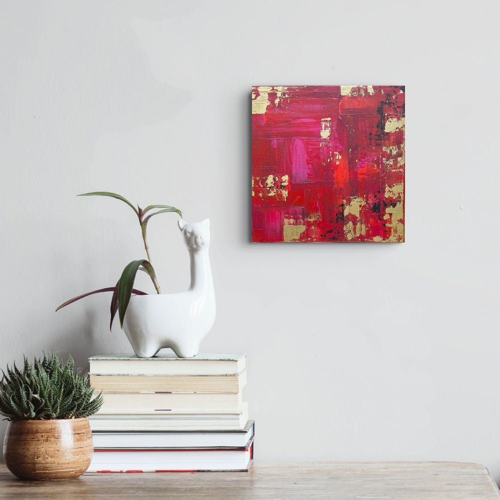 A square abstract canvas painting with bold red, pink, and gold brushstrokes hangs on a light gray wall above a stack of books and a white ceramic llama planter with a green plant. Samhain No.1 Original Canvas Painting by Julie Bevan Art