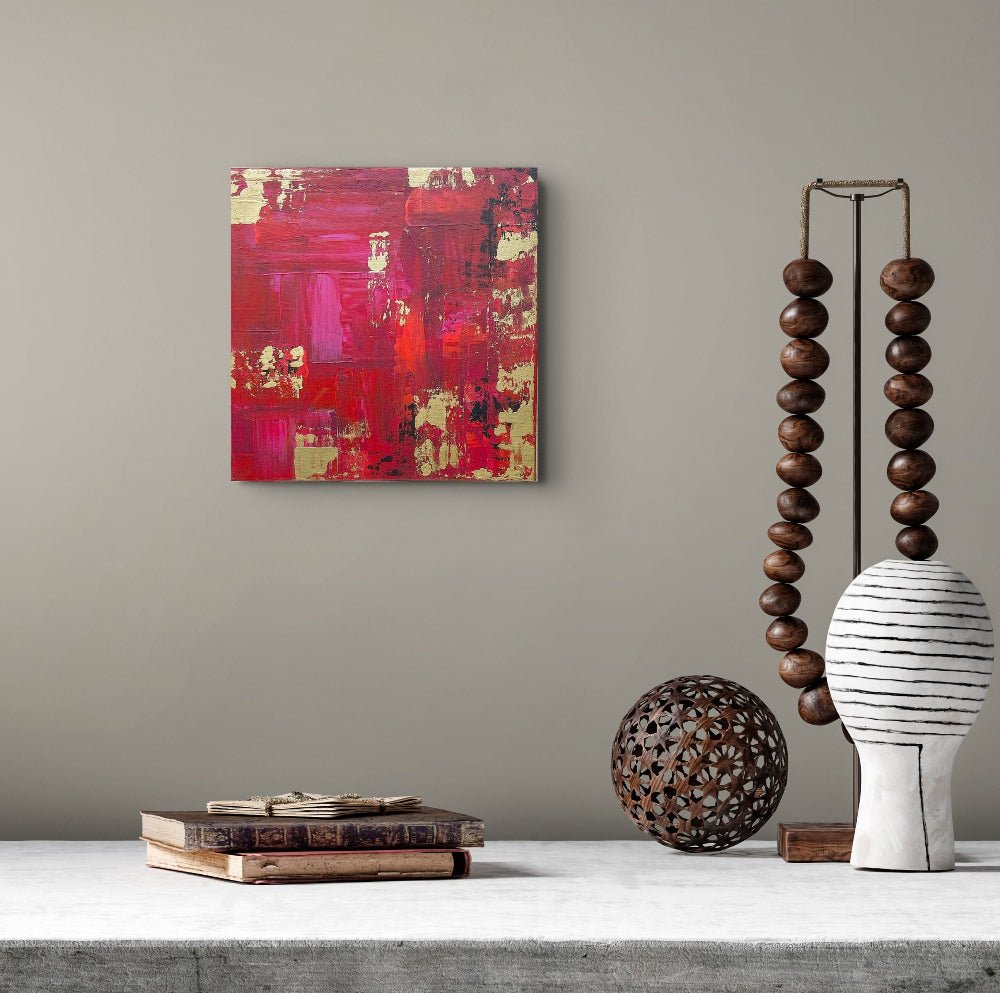 Abstract canvas wall art featuring bold red, magenta, and gold brushstrokes, displayed on a neutral gray wall above a styled tabletop with books, a wooden bead sculpture, and a white ceramic vase. Samhain No.1 Original Canvas Painting by Julie Bevan Art