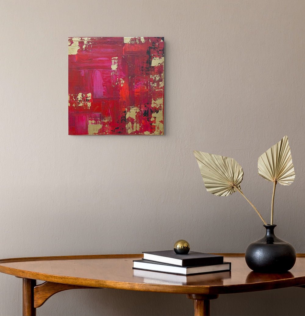 Abstract red and pink canvas wall art hanging on a beige wall above a wooden console table with books and a black vase holding dried palm leaves. Samhain No.1 Original Canvas Painting by Julie Bevan Art