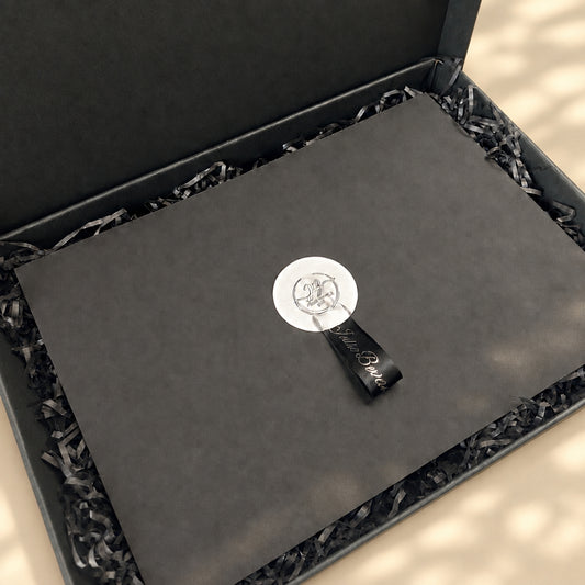 A black presentation box with shredded black paper filler, featuring a matte black insert card with a circular silver logo seal and a small black branded tag.