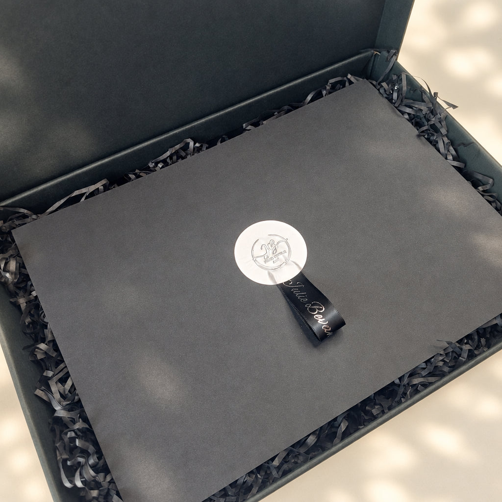 A black presentation box with shredded black paper filler, featuring a matte black insert card with a circular silver logo seal and a small black branded tag.
