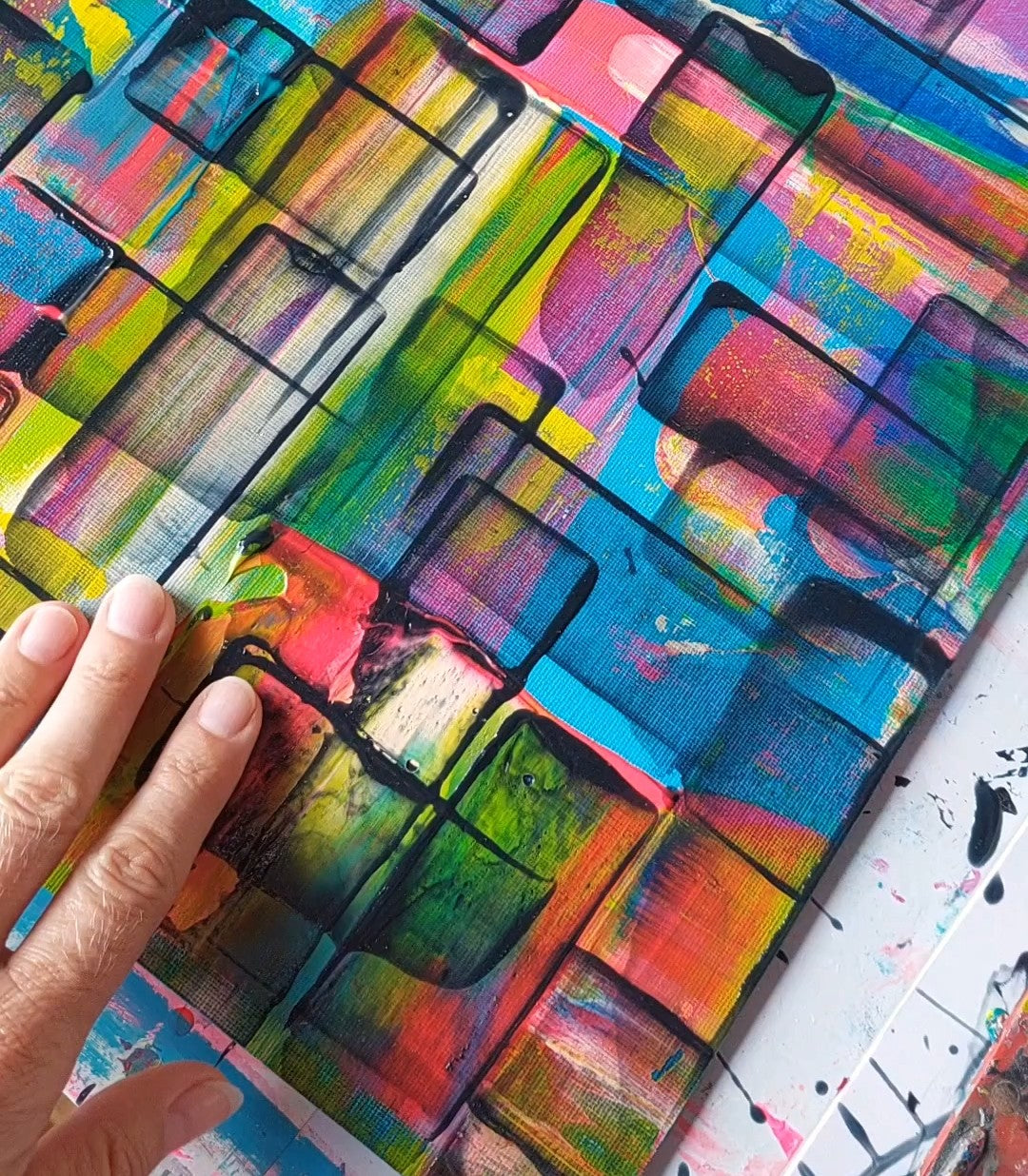 A close-up of a colorful abstract painting with bold brushstrokes in blue, green, yellow, pink, orange, and black, with a hand touching the canvas.