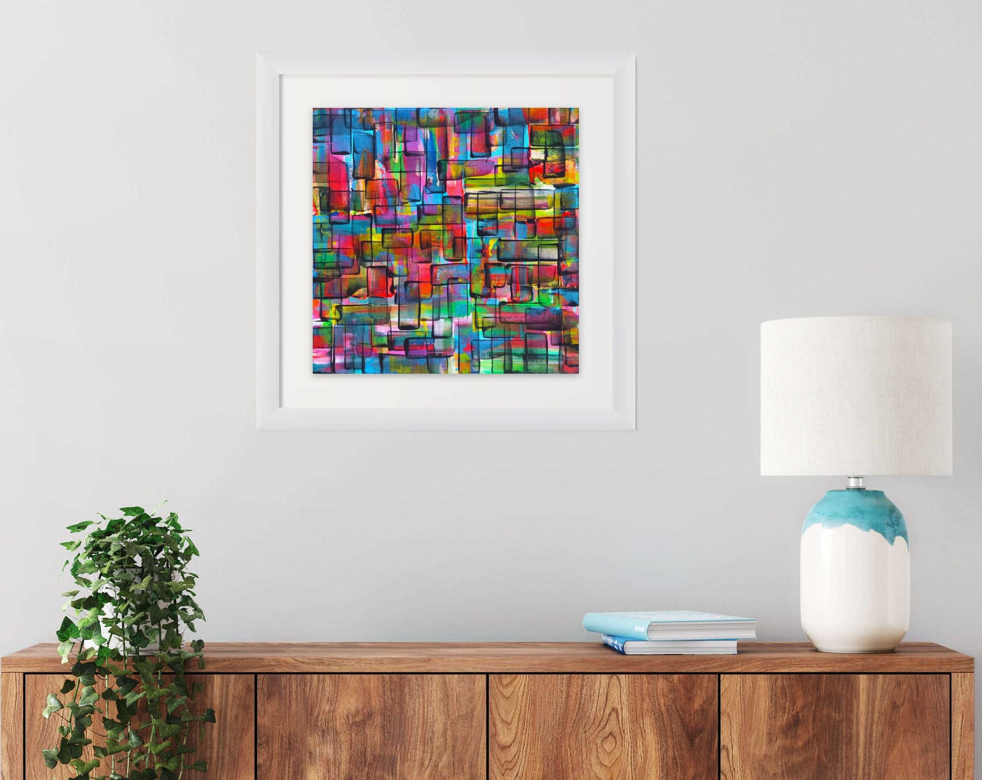 A framed abstract geometric art print with a colorful mosaic pattern hangs on a light gray wall above a wooden sideboard. The print is enclosed in a white frame and is styled in a home interior setting with a potted plant, books, and a lamp on the cabinet.