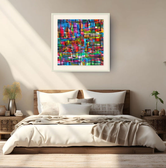 A colorful abstract framed wall art print hangs above a neatly made bed in a modern, sunlit bedroom. The artwork features a vibrant mosaic of red, blue, green, yellow, and purple geometric shapes inside a white frame. Refraction limited edition print by Julie Bevan