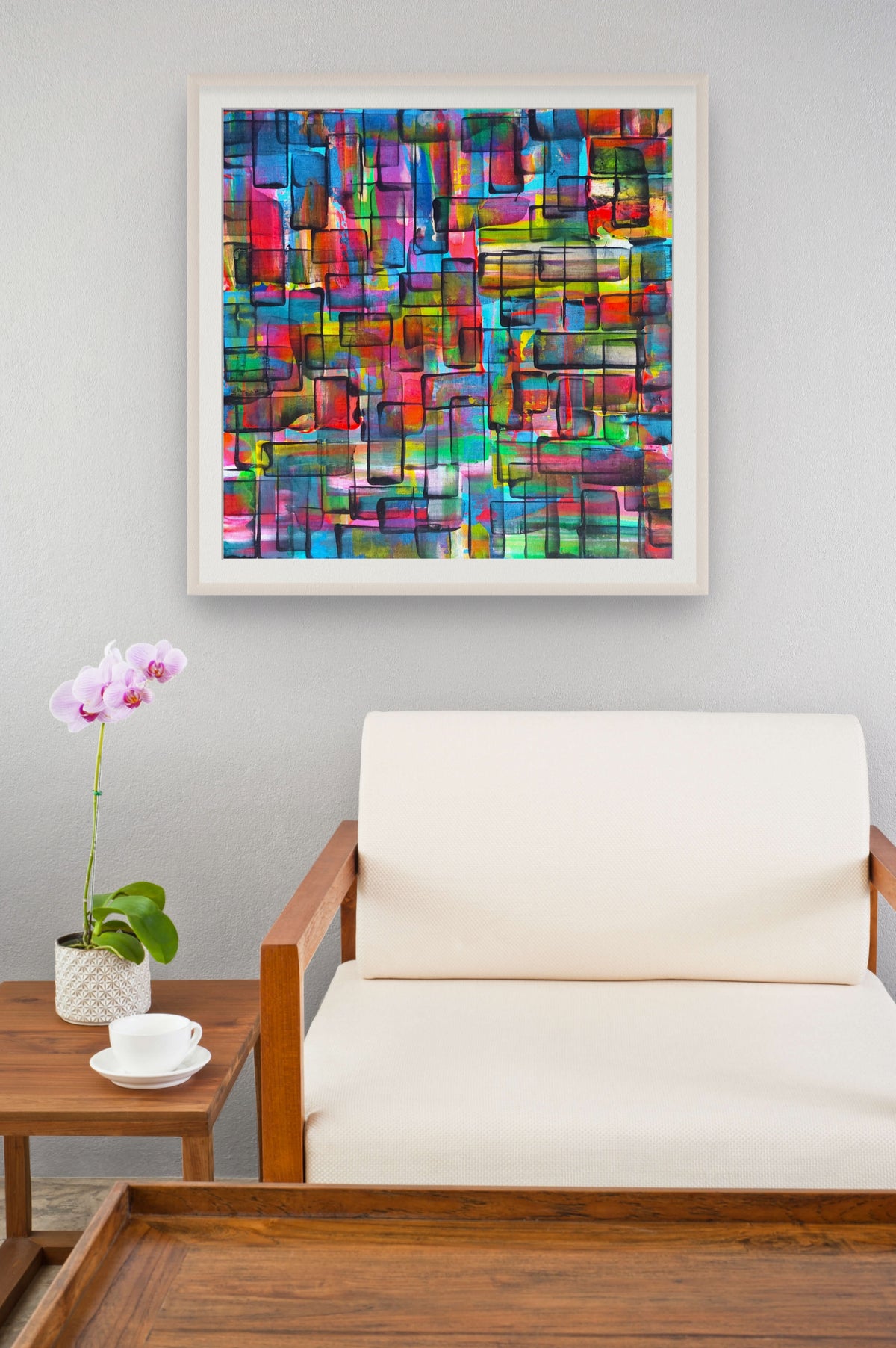 A colorful abstract geometric wall art print in a white frame, displayed on a light gray wall above a cream-colored armchair with a wooden side table holding a pink orchid in the foreground. Refraction limited edition print by Julie Bevan
