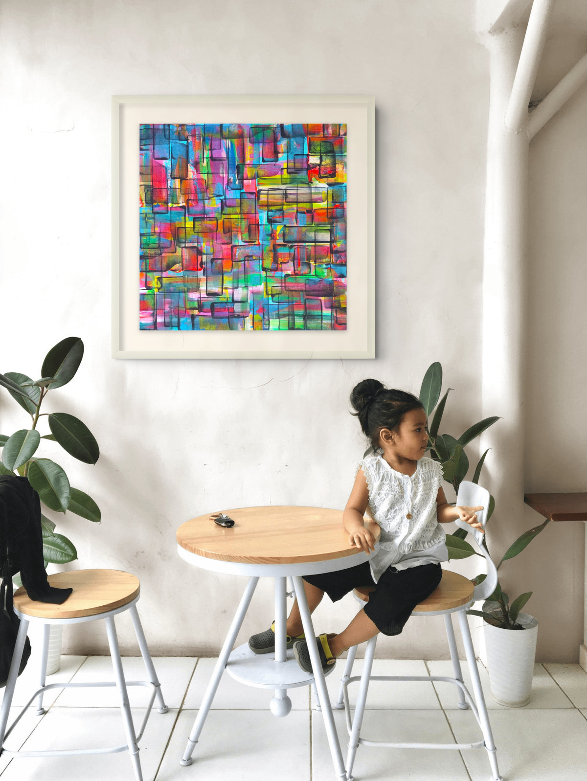 A colorful abstract geometric wall art print with a white frame hangs on a white wall above a small round wooden table. A young girl sits on a white stool at the table, and potted plants are visible in the room. Refraction limited edition print by Julie Bevan