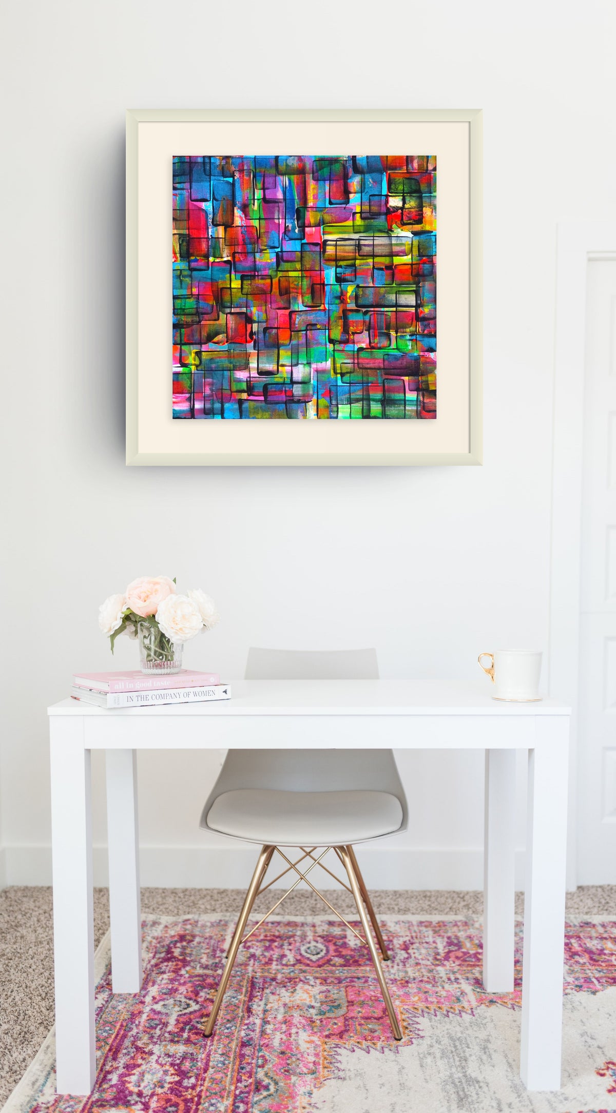 A framed abstract geometric art print with a vibrant multicolor mosaic pattern, displayed on a white wall above a modern white desk and gray chair in a bright room. Refraction limited edition print by Julie Bevan
