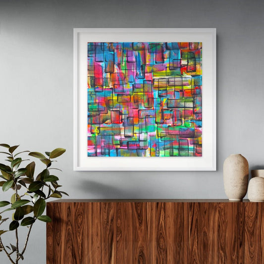 A framed abstract geometric art print with a colorful mosaic pattern in shades of blue, pink, red, yellow, green, and orange, displayed on a gray wall above a wooden console table with decorative vases and a plant. Refraction limited edition print by Julie Bevan