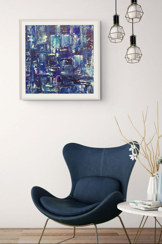 A framed abstract painting with deep blue, purple, and black brushstrokes hangs on a white wall above a modern navy blue armchair in a styled living room setting. Samhain No. 2 Limited Edition Print by Julie Bevan