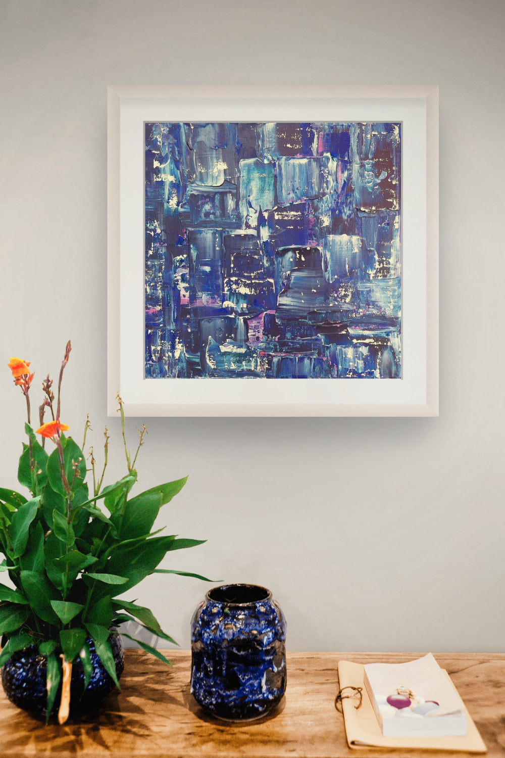 A framed abstract blue painting hangs on a light gray wall above a wooden table with a green plant in a blue vase and a small blue decorative jar. Samhain No. 2 Limited Edition Print by Julie Bevan