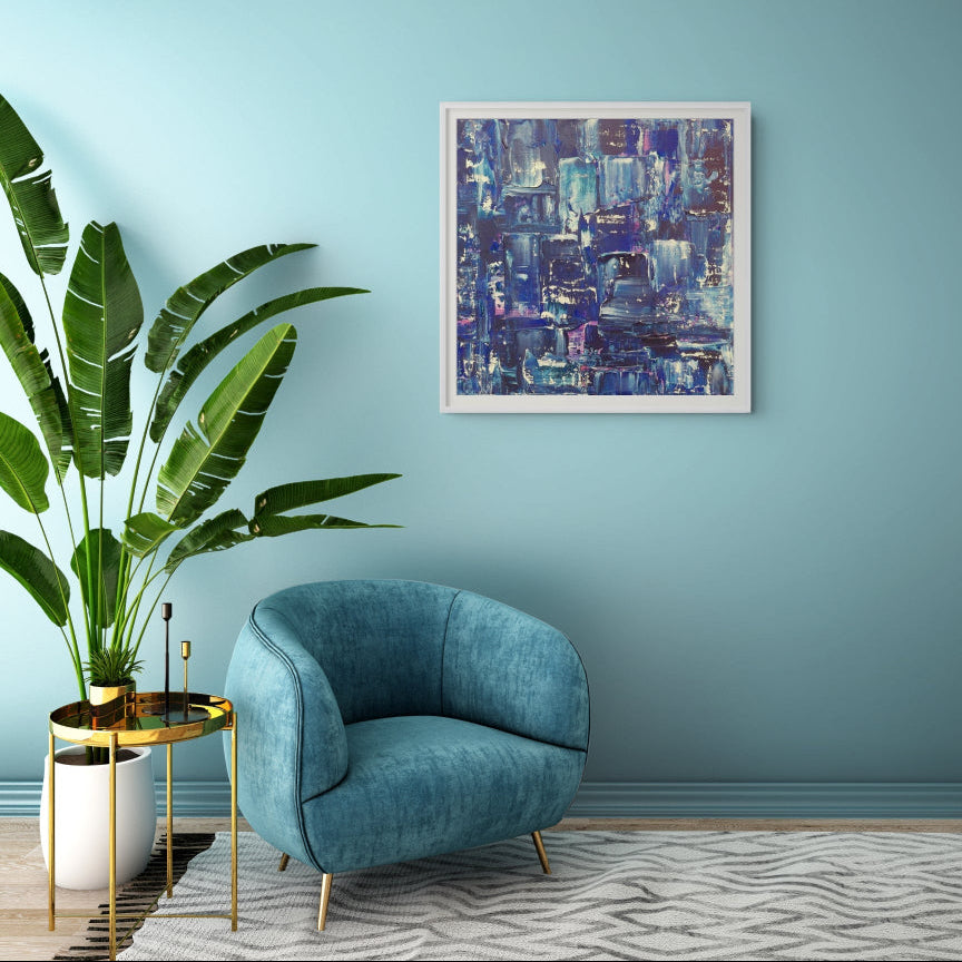 A square framed abstract painting with blue, purple, and white brushstrokes hangs on a light blue wall in a styled room with a teal velvet armchair, a potted green plant, and a patterned rug. Samhain No. 2 Limited Edition Print by Julie Bevan