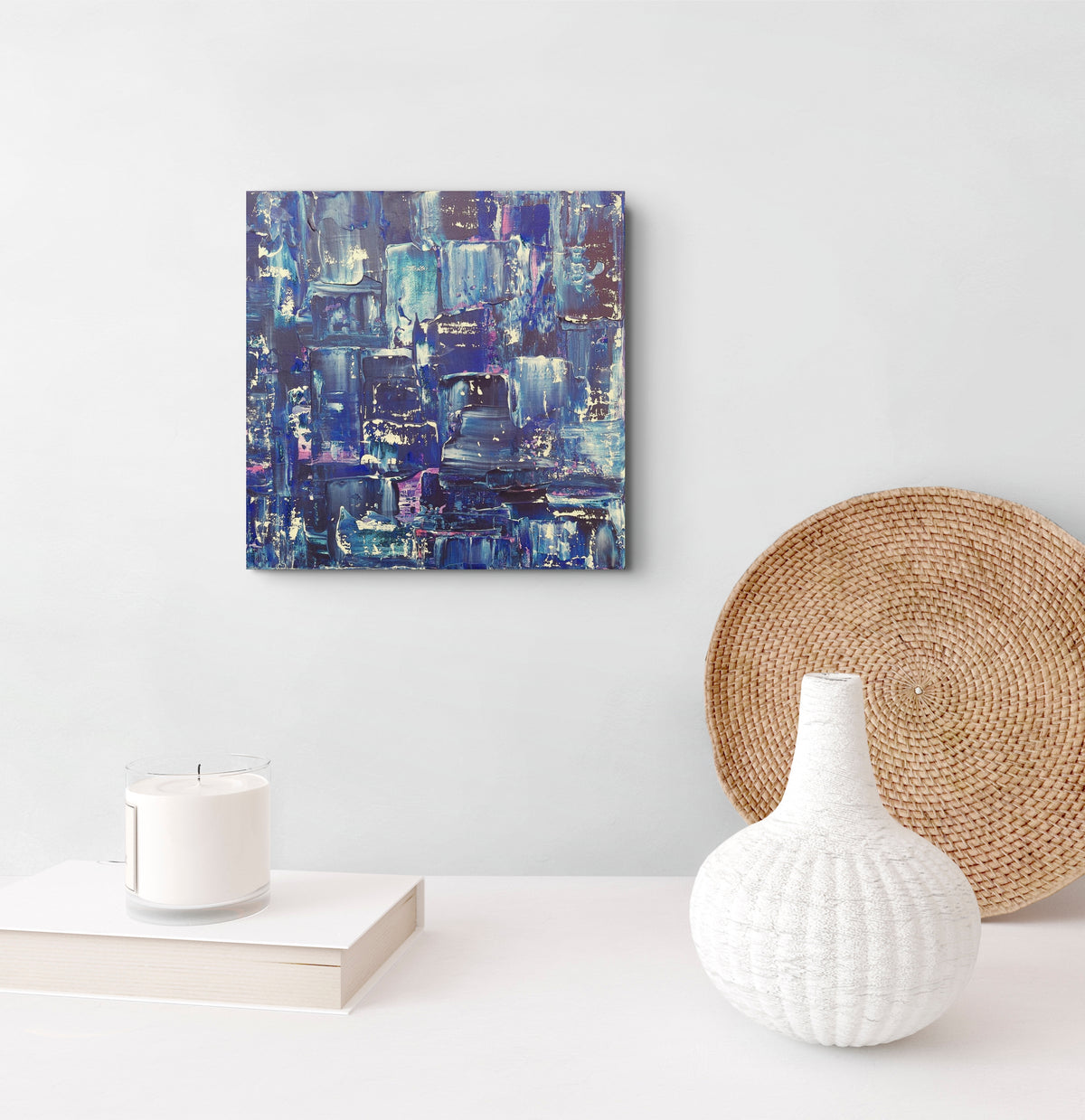 An abstract canvas painting featuring shades of blue, purple, and white, displayed on a light gray wall above a white table styled with a white vase, a woven round mat, and a white candle. Samhain No.2 Original Canvas Painting by Julie Bevan