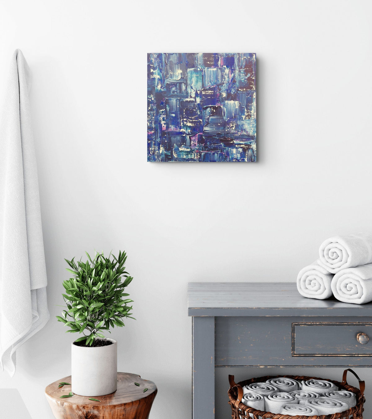A square abstract canvas print with blue, purple, and white brushstrokes hangs on a white wall in a styled bathroom setting. Below the canvas, a gray wooden vanity table holds three rolled white towels, and to the left, a small potted green plant sits on a wooden stool next to a white towel hanging on the wall. Samhain No.2 Original Canvas Painting by Julie Bevan