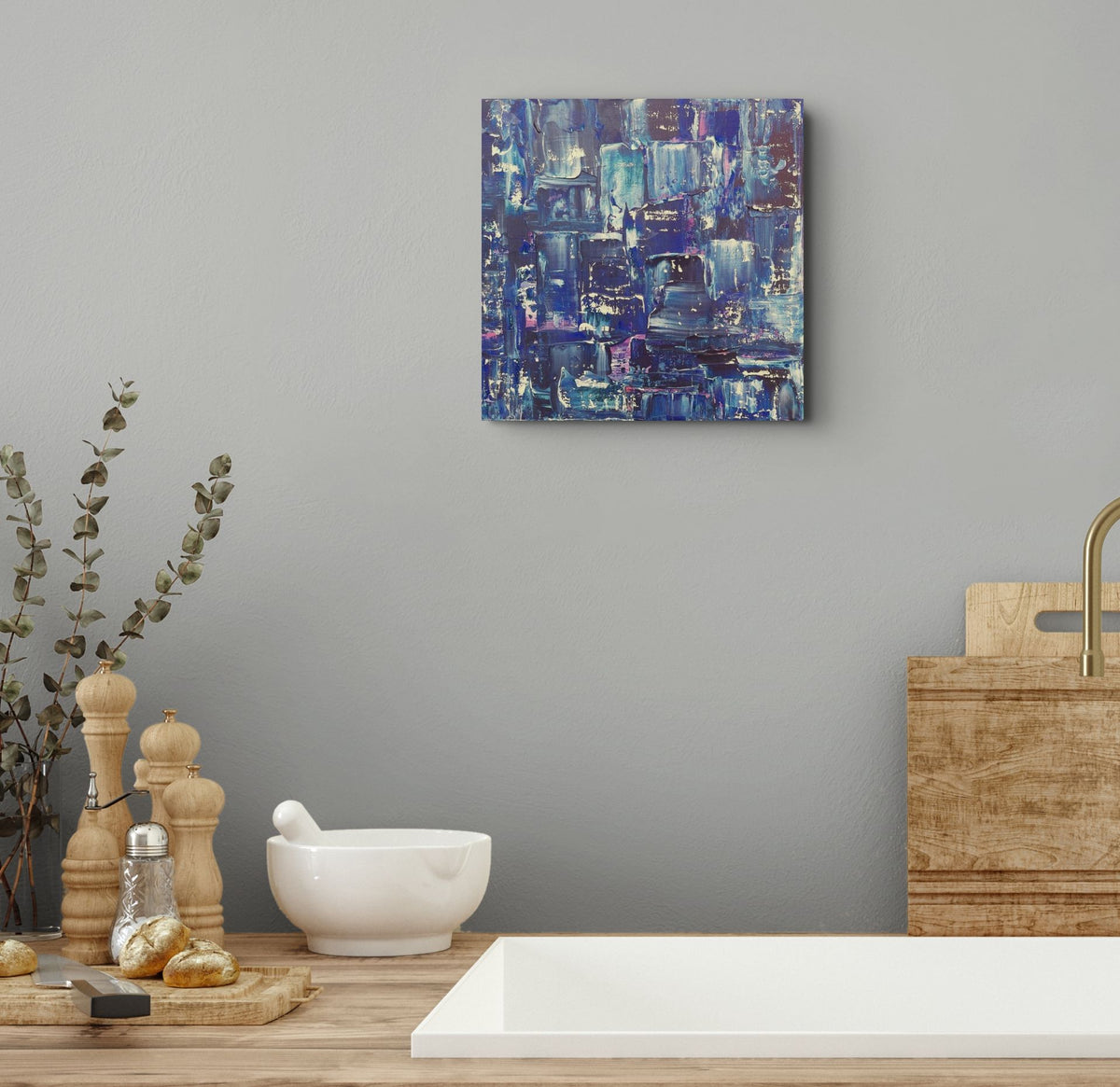 A square abstract canvas print with blue, purple, and white brushstrokes hangs on a gray wall above a wooden countertop with a white bowl, wooden peeler, and other kitchen props. Samhain No.2 Original Canvas Painting by Julie Bevan
