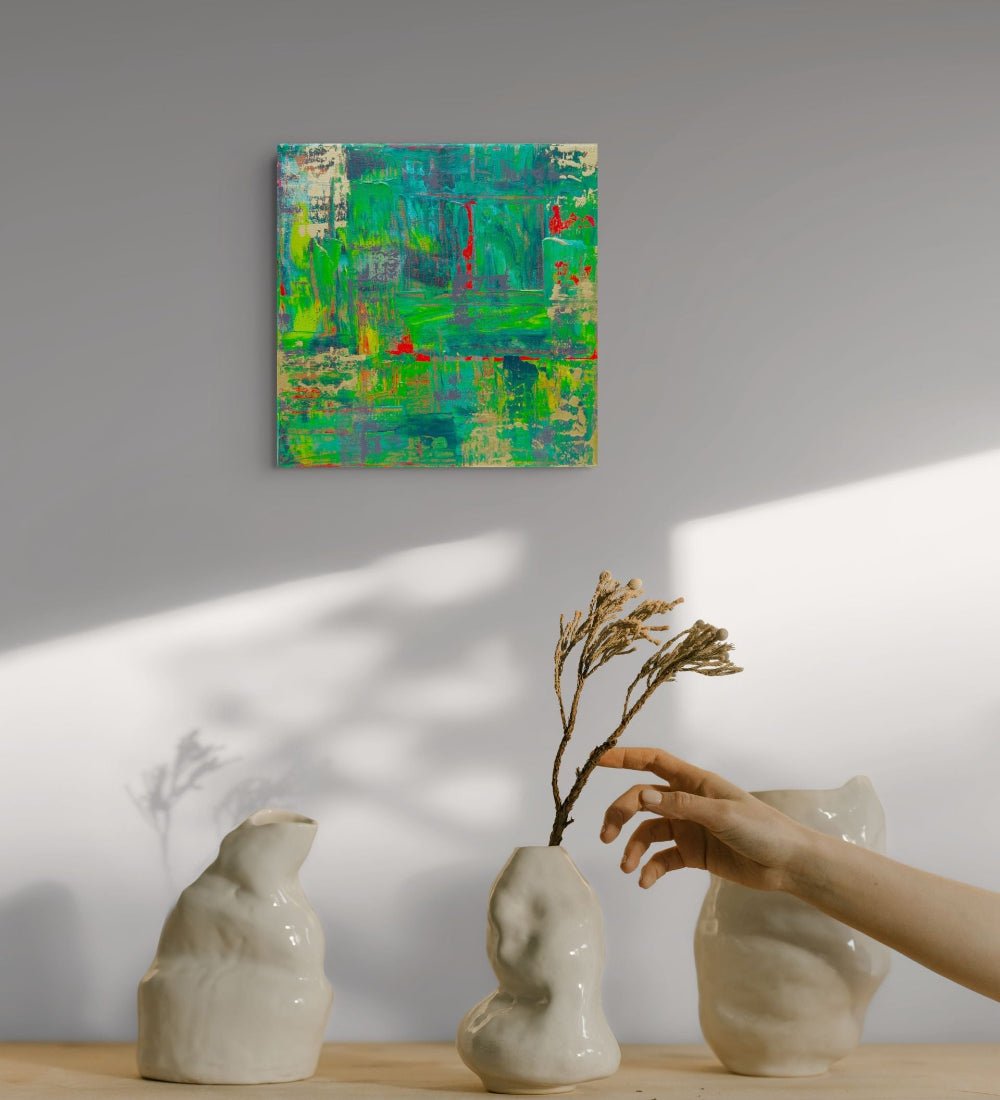 An abstract painting with vibrant green, blue, and red brushstrokes hangs on a light gray wall above a wooden table with three textured off-white ceramic vases and a hand holding a dried twig. samhain no 3 original canvas art by julie bevan