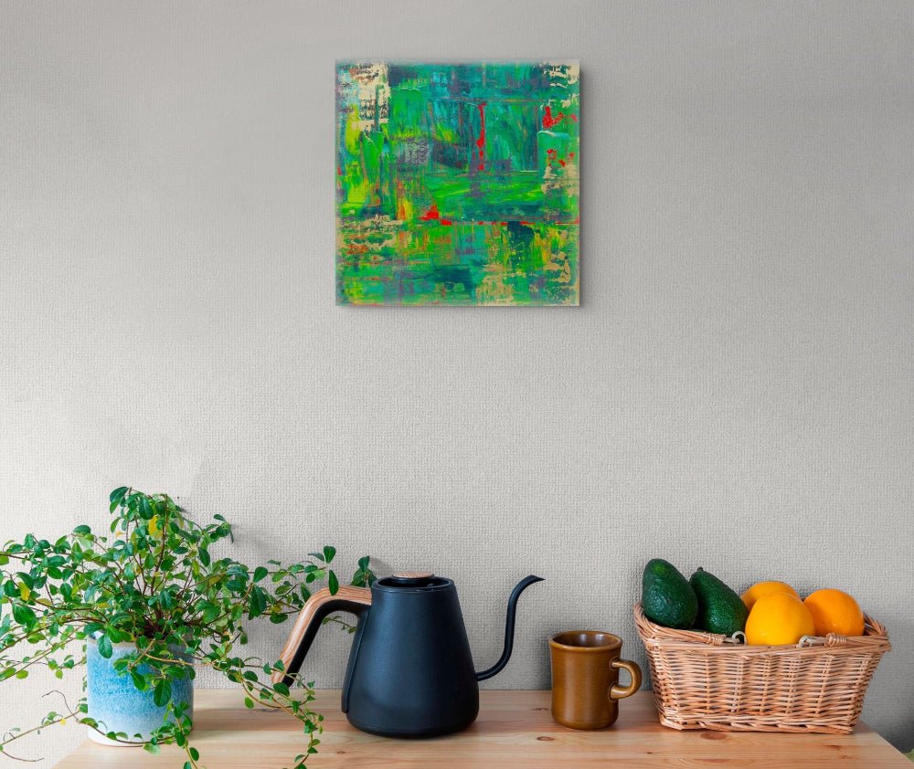 An abstract painting with green, blue, and red brushstrokes hangs on a light gray wall above a wooden table styled with a potted plant, a matte black gooseneck kettle, a brown mug, and a wicker basket holding avocados and oranges. samhain no 3 original canvas art by julie bevan
