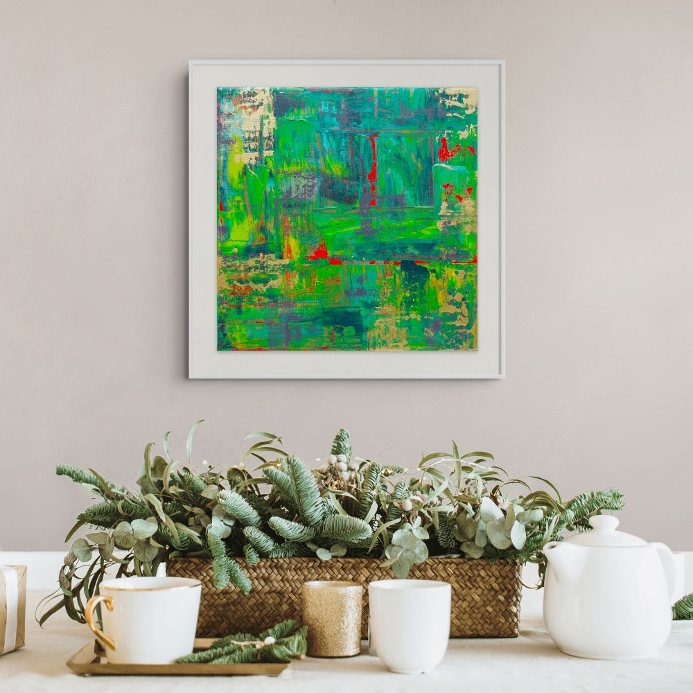 A framed abstract painting with vibrant green, teal, and red brushstrokes hangs on a light beige wall above a styled tabletop with potted greenery, white mugs, and a white teapot. Samhain no. 3 limited edition print by Julie Bevan