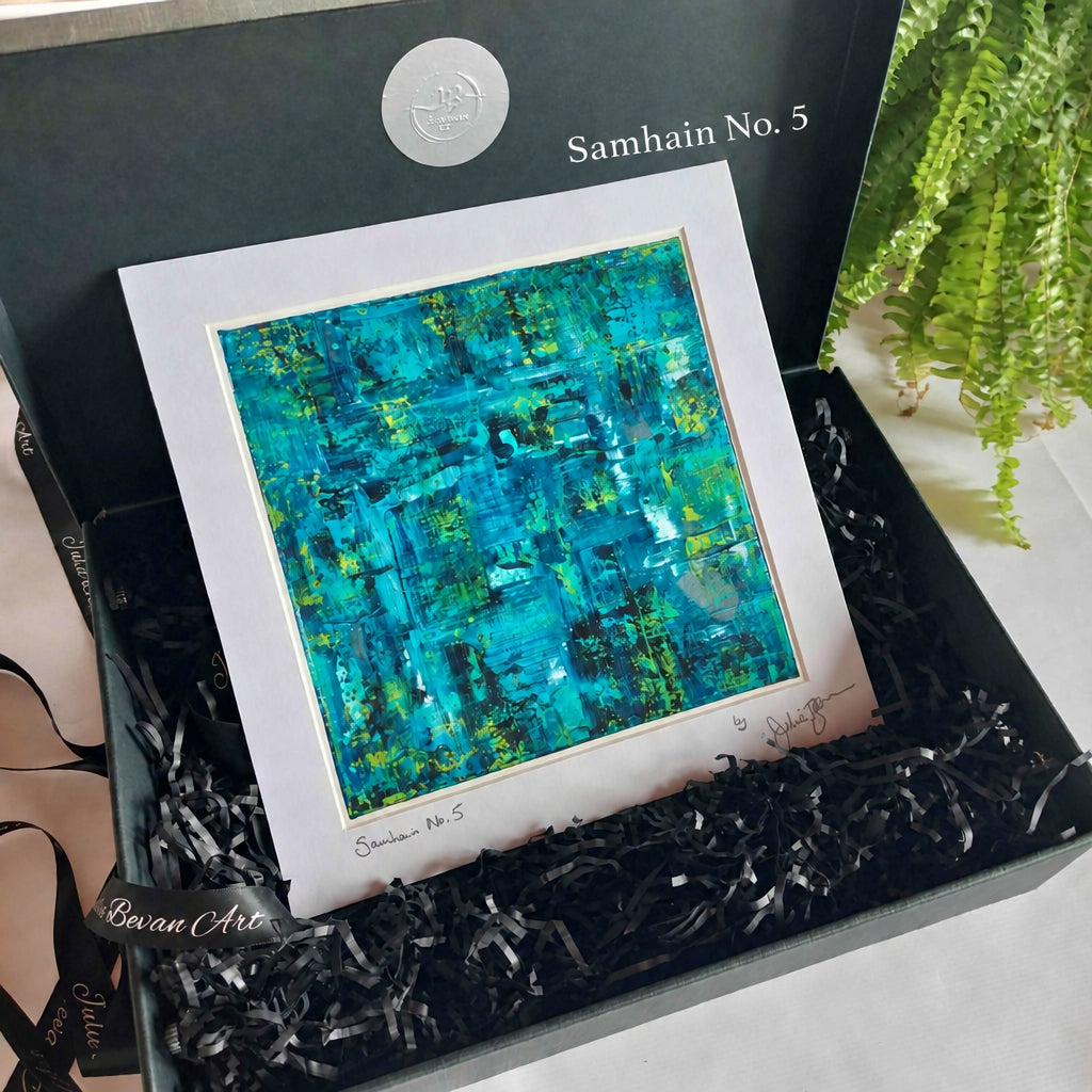 An abstract art print featuring vibrant turquoise, teal, and green brushstrokes, matted with a wide white border and presented inside a black gift box with black shredded paper filler. The box lid has a silver circular logo, and a green fern is visible in the background.