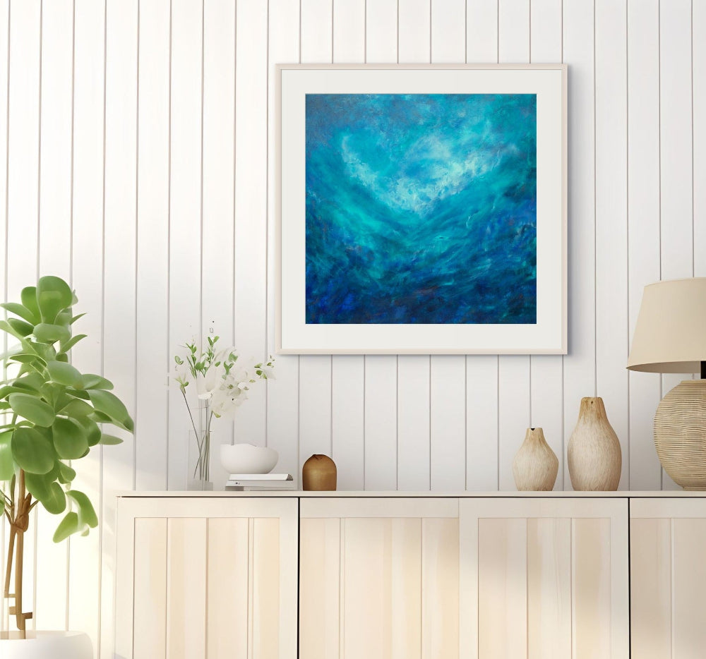 A framed abstract painting featuring swirling shades of blue and teal, displayed on a white paneled wall above a light wood console table with decorative vases, a plant, and a lamp.