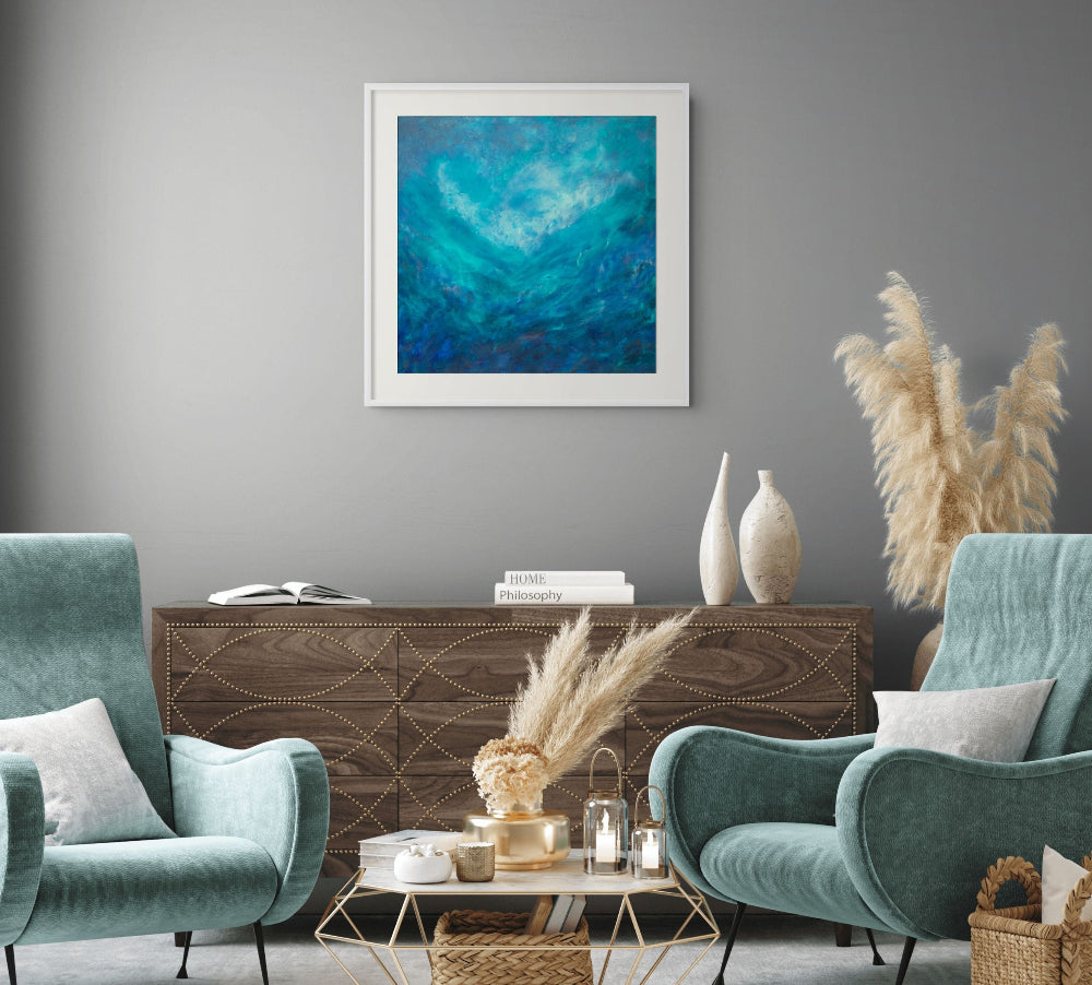 A framed abstract painting with swirling blue and teal tones hangs on a gray wall above a wooden console in a styled living room with two teal armchairs, a coffee table, and pampas grass.