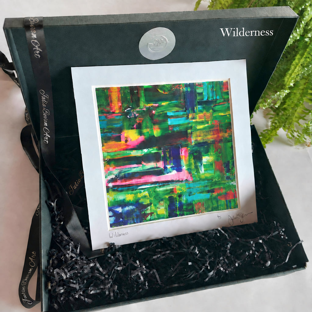 An abstract colorful painting presented inside a black gift box with shredded black paper filler and a branded ribbon. The artwork features bold brushstrokes in green, blue, pink, orange, and yellow on a white mat board.